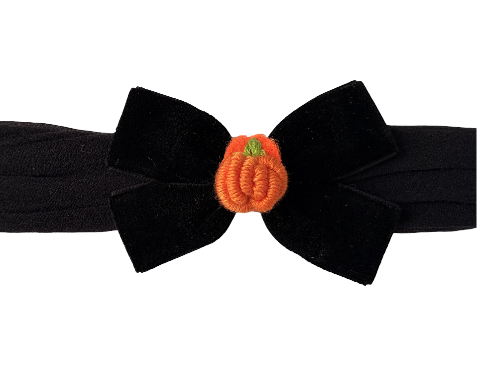A black fabric headband with a large bow in the center, decorated with a small orange pumpkin embellishment featuring a tiny green stem, shown against a plain white background.