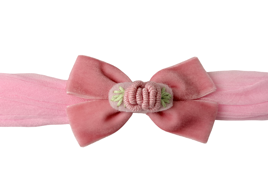 A pink velvet bow headband with a central decorative knot and small green embroidered details, shown against a plain white background.