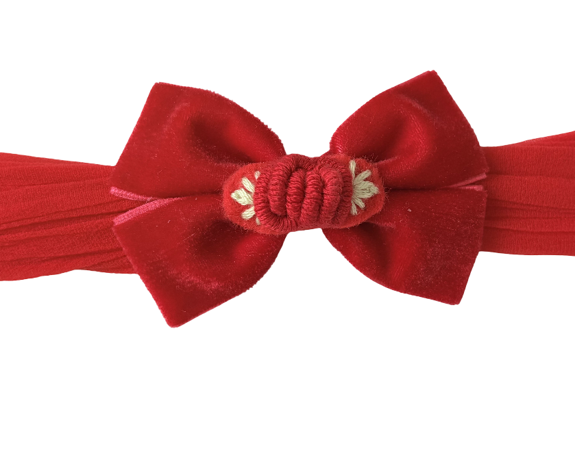 A red velvet bow headband with a decorative knotted center and small white floral embellishments, shown against a plain white background.
