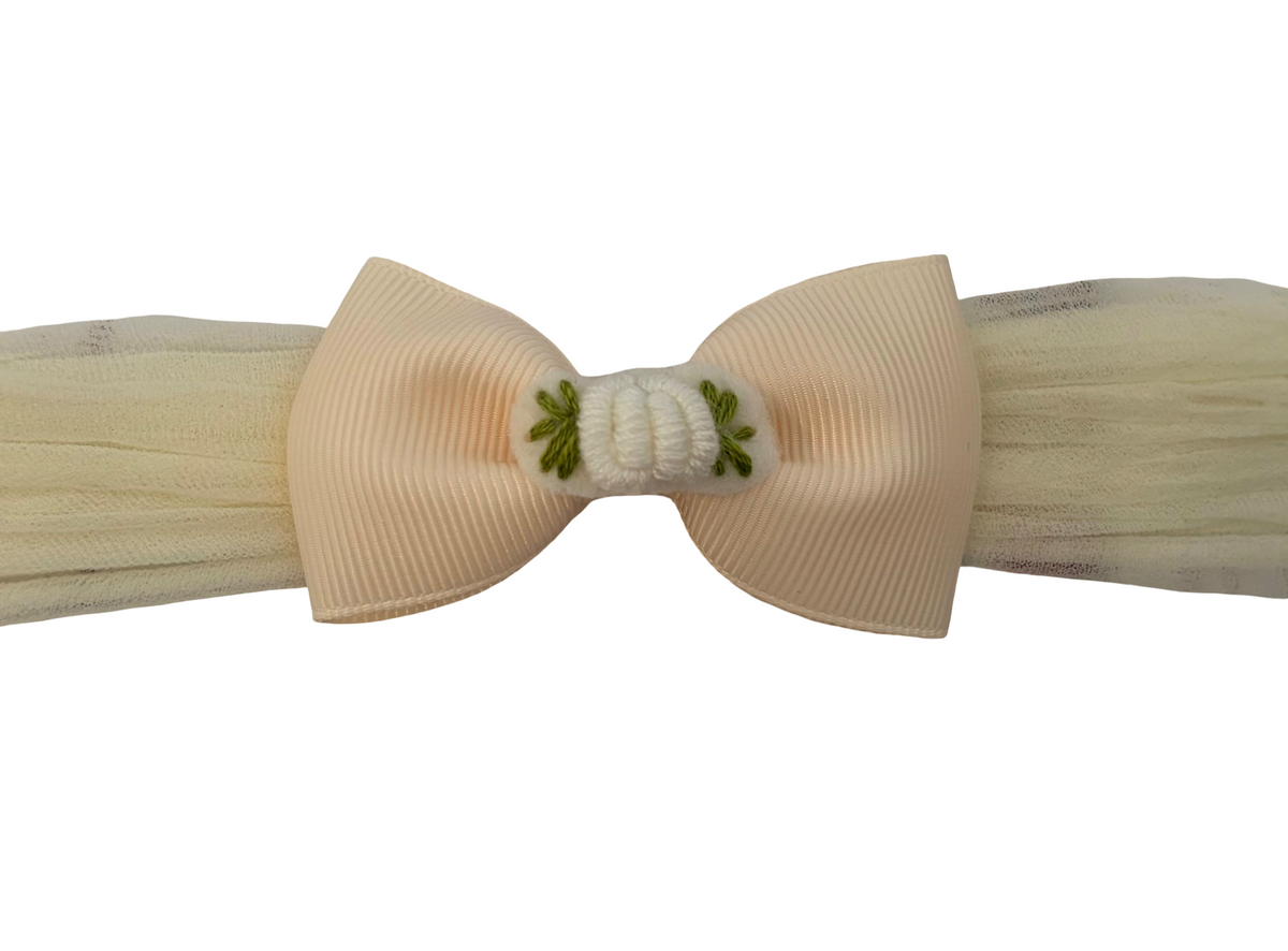 A cream-colored grosgrain ribbon bow with a white knotted center and small green embroidered leaves, displayed on a matching cream tulle band against a white background.