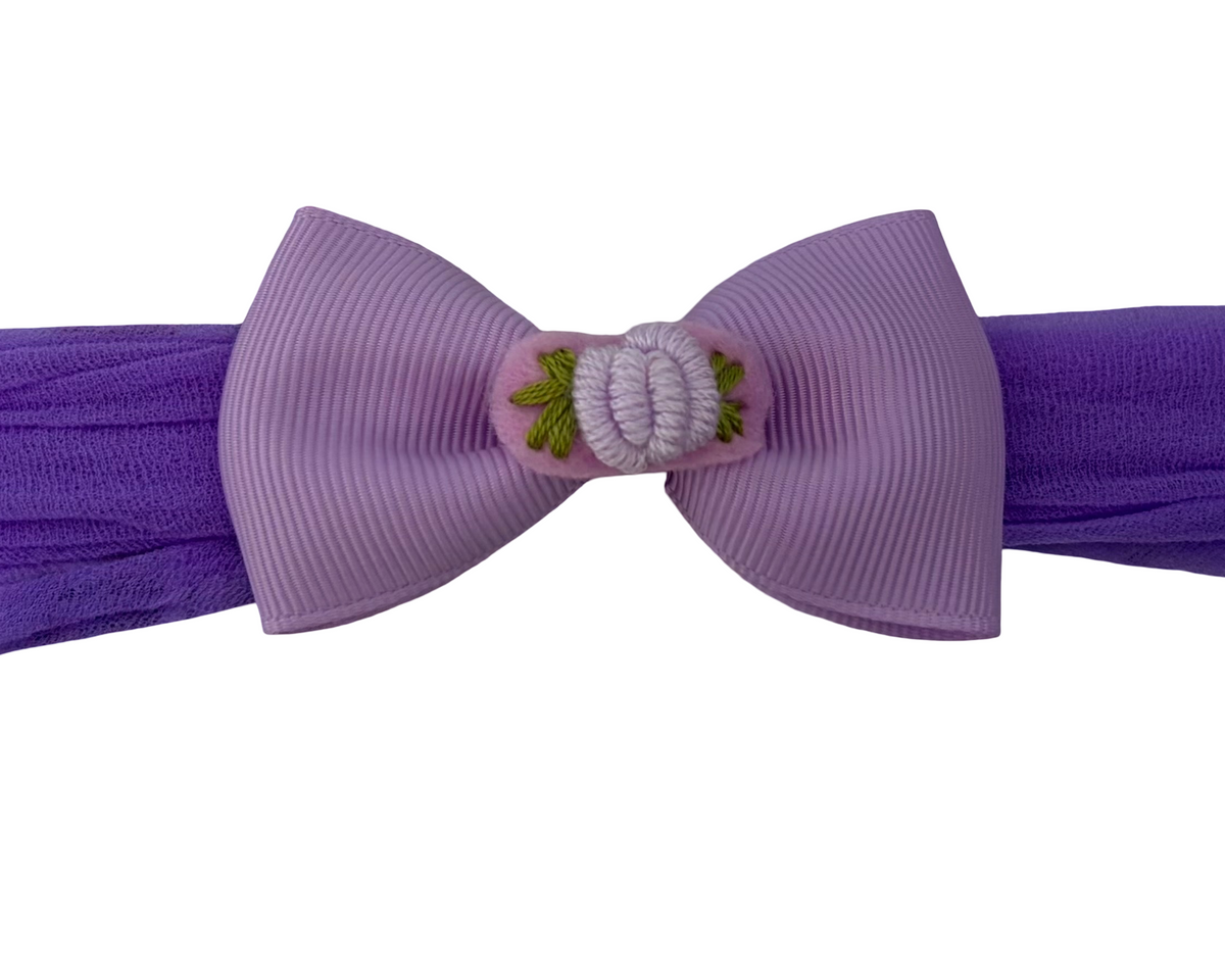 A purple grosgrain ribbon bow with a central decorative knot and small green leaf embroidery, attached to a matching purple ribbon headband, shown on a plain white background.