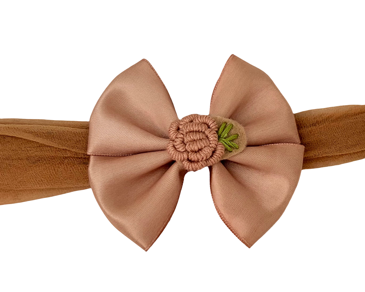 A dusty rose satin bow headband with a textured brown elastic band and a small green embroidered accent in the center, shown against a plain white background.