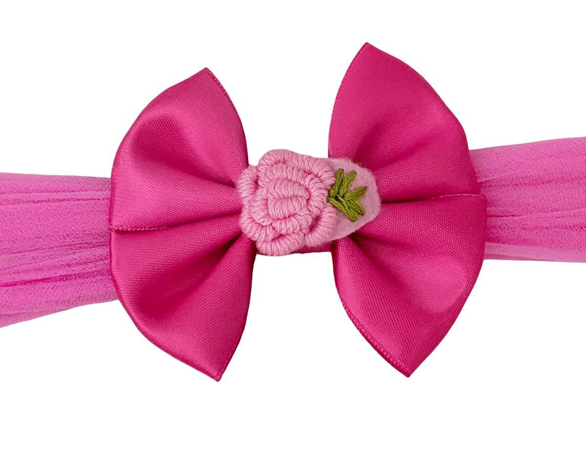 A bright pink fabric headband with a large bow in the center, decorated with a small pink crocheted flower and green leaf accent.